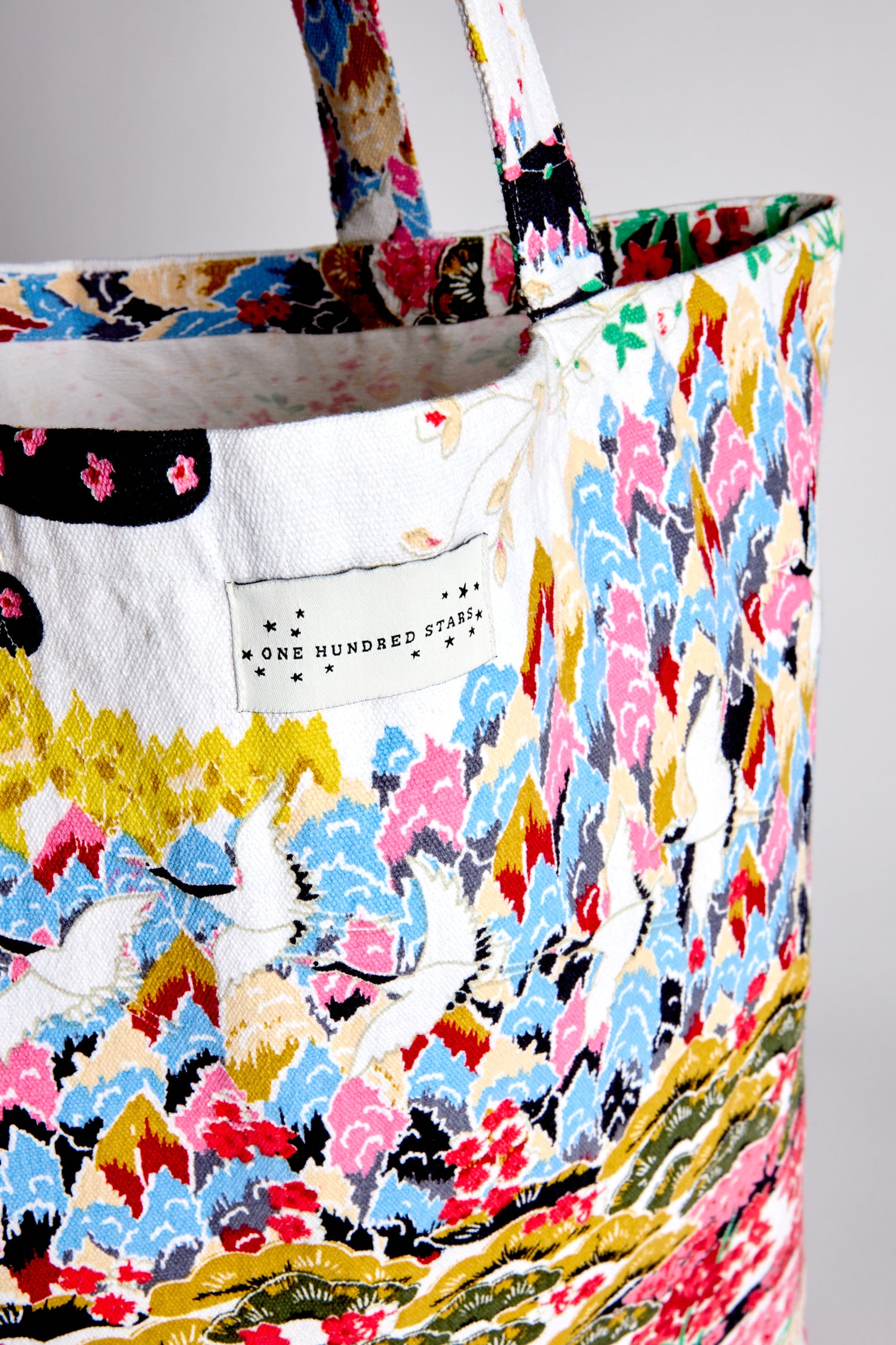 Close-up of a colorful Spring Print Canvas Bag featuring a floral and leaf pattern made from sustainable cotton. The label reads "One Hundred Stars." This reusable shopping bag is ideal for eco-conscious fashion enthusiasts.