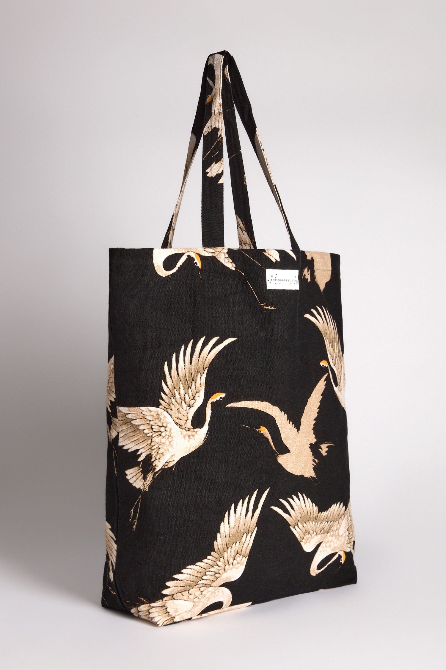 The Stork Canvas Bag is a black tote with a cream crane pattern, made from sustainable cotton and featuring dual handles on a plain background.