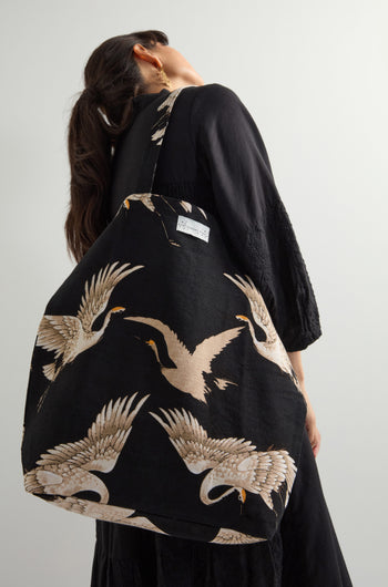 A dark-haired woman in a black dress holds the eco-friendly Stork Canvas Bag, made from sustainable cotton and featuring a striking crane design.