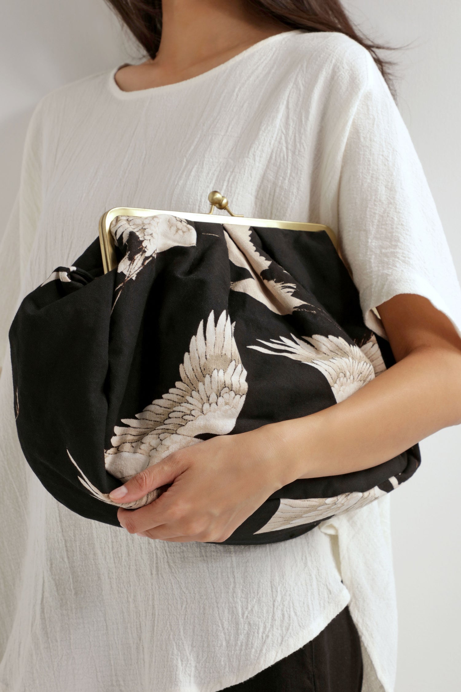 A person in a white top holds the Stork Print Clasp Bag—crafted from organic cotton, this stylish accessory features a crane bird motif, art deco design elements, and a bold metal clasp frame.