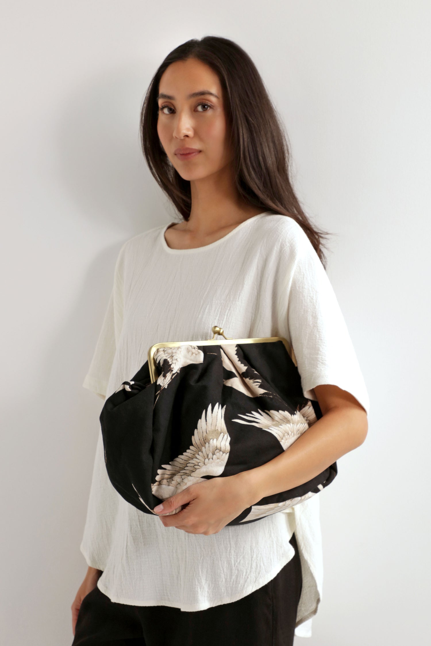 A woman stands against a white wall, wearing a loose white top and carrying the Stork Print Clasp Bag made from organic cotton under her arm.
