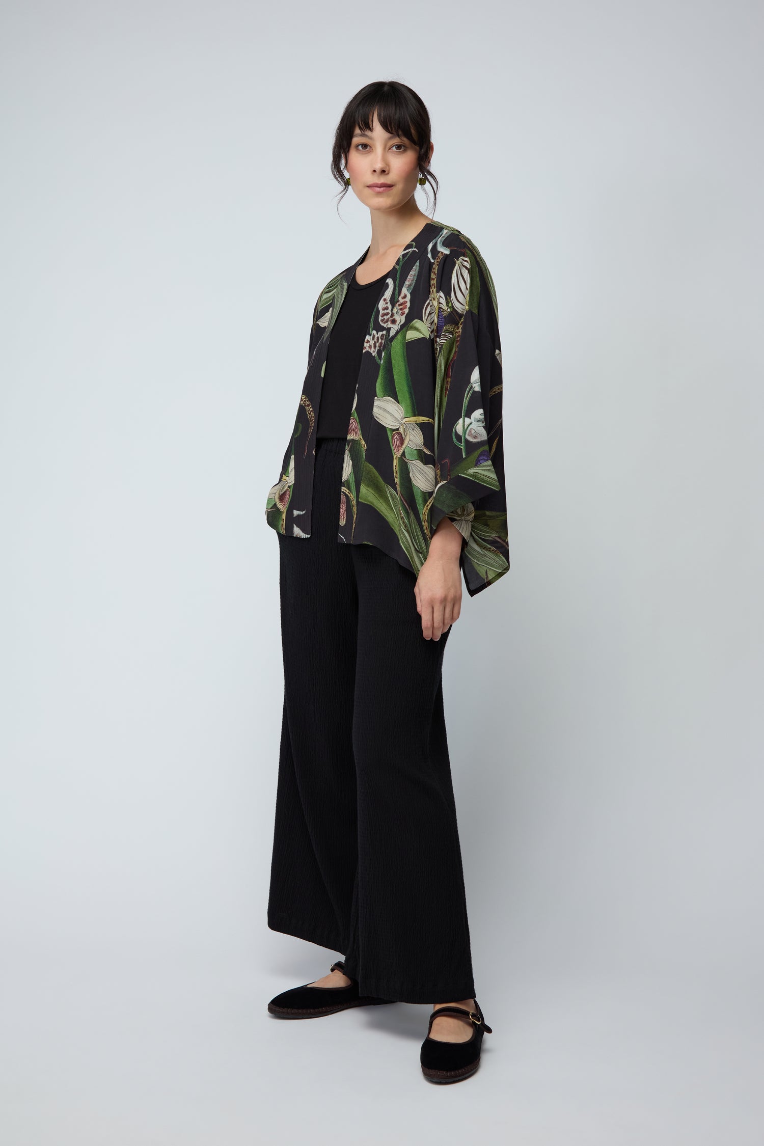 A woman stands against a plain background in wide-leg black pants and the Orchid Crepe Kimono, a loose top crafted from sustainable fabric featuring a delicate botanical print.