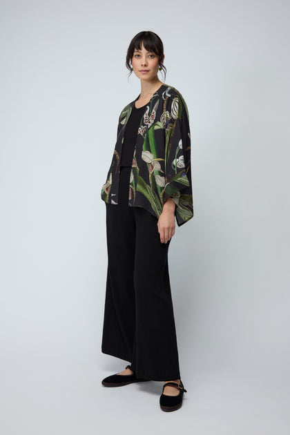 A woman stands against a plain background in wide-leg black pants and the Orchid Crepe Kimono, a loose top crafted from sustainable fabric featuring a delicate botanical print.