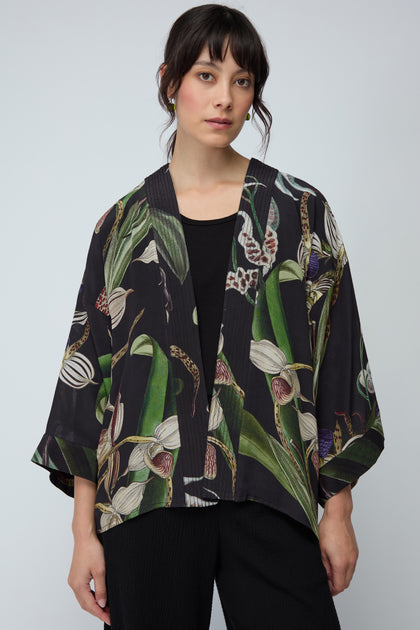 A woman models the Orchid Crepe Kimono—featuring a black botanical print—over a black top and pants, set against a plain light background.