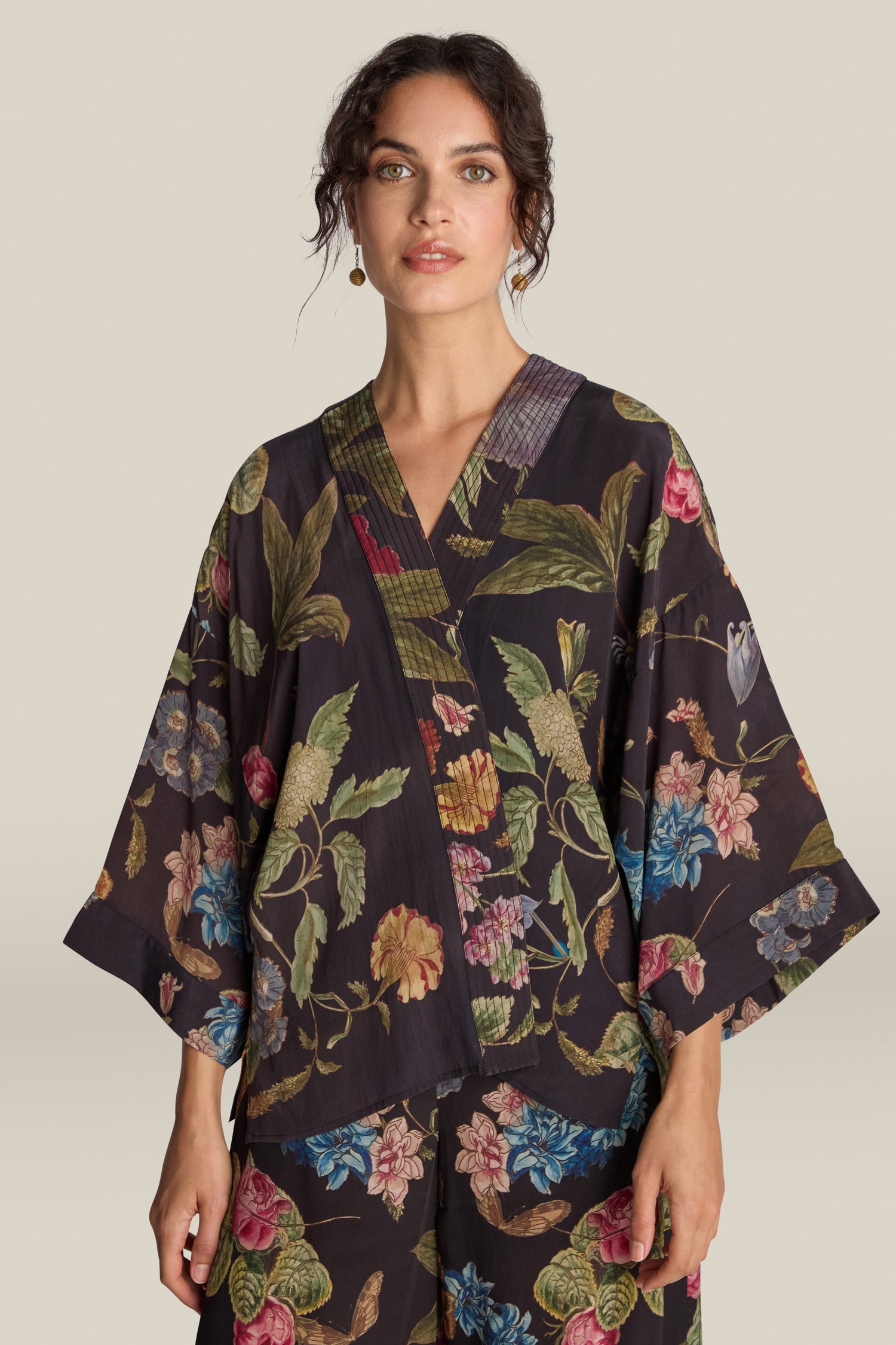 A woman with dark hair in a partly pinned-up style, wearing the Primula Crepe Kimono featuring a dark floral print top and pants, stands against a plain beige background.