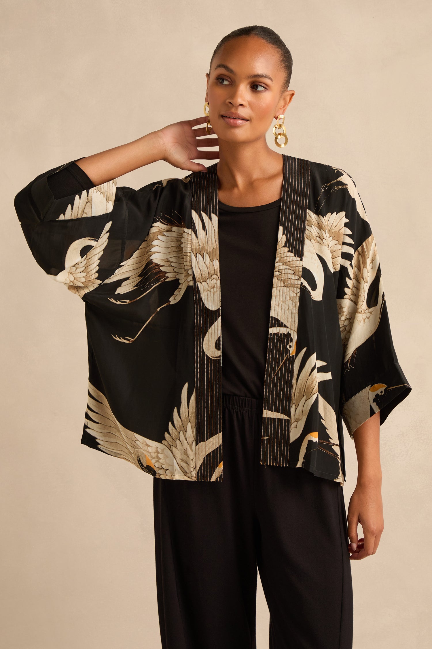 A woman wearing the Stork Crepe Kimono in black with a white crane pattern, paired with a black top and black pants, stands against a beige background.