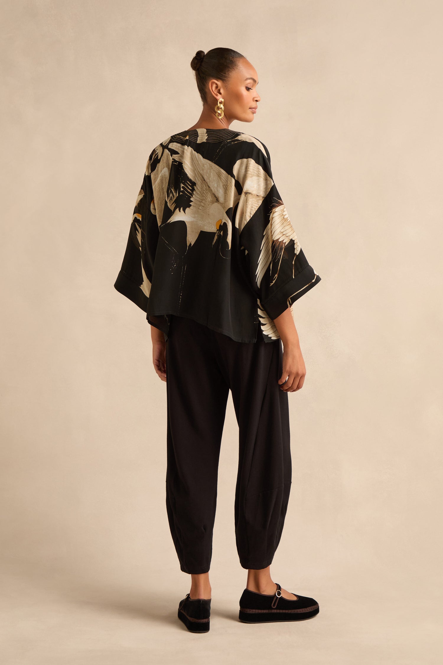 A person is standing with their back to the camera, wearing the Stork Crepe Kimono—a black Art Deco-inspired top with an exclusive stork print—paired with black pants, black shoes, and gold earrings against a beige background.