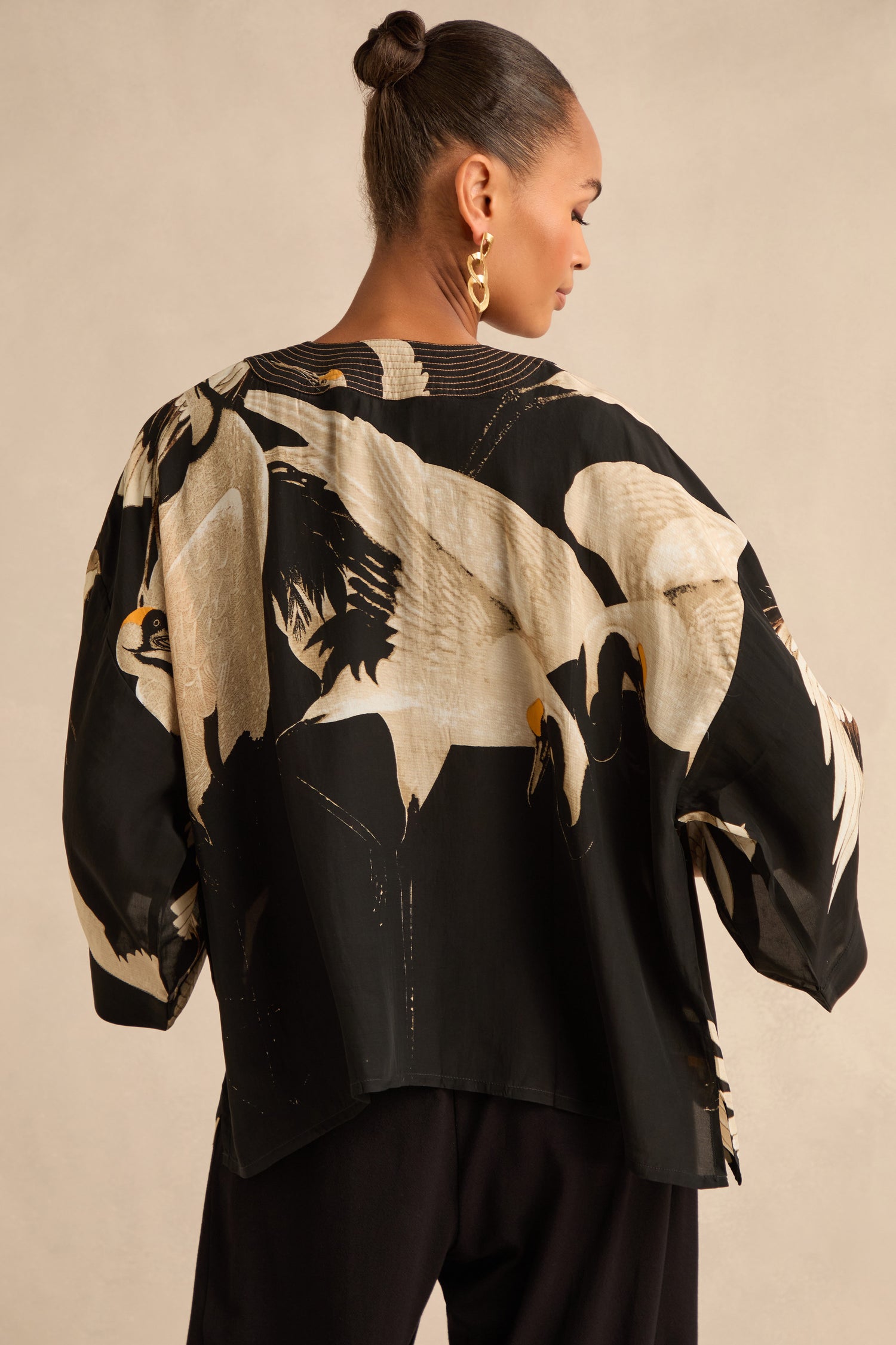 A woman, seen from behind, wears the Stork Crepe Kimono—a black kimono-style top featuring a subtle Art Deco print and intricate white crane detailing—paired with black pants and gold hoop earrings; her hair is styled in a bun.