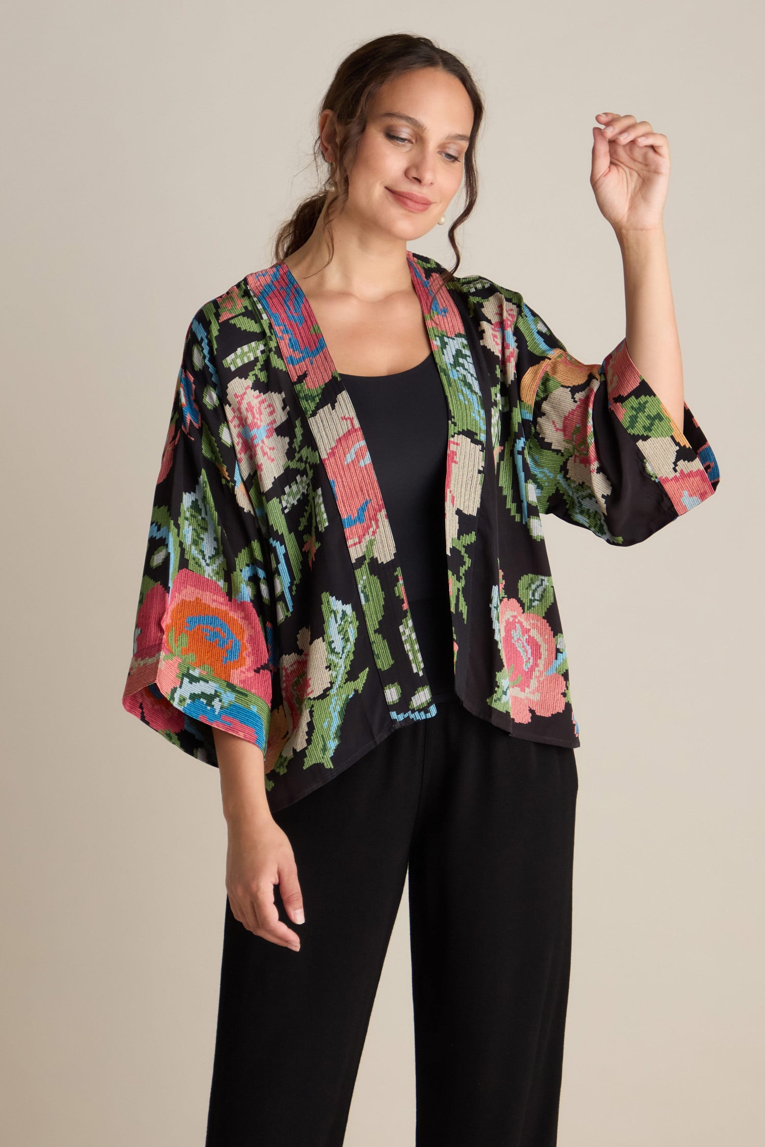 A woman dons the Woven Flower Crepe Kimono over a black top and matching pants. Standing against a plain background, she closes her eyes and raises one arm gracefully.