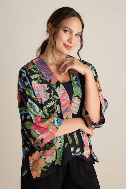 A person with a slight smile poses in a brightly colored Woven Flower Crepe Kimono featuring a floral motif, layered perfectly over a plain dark shirt.