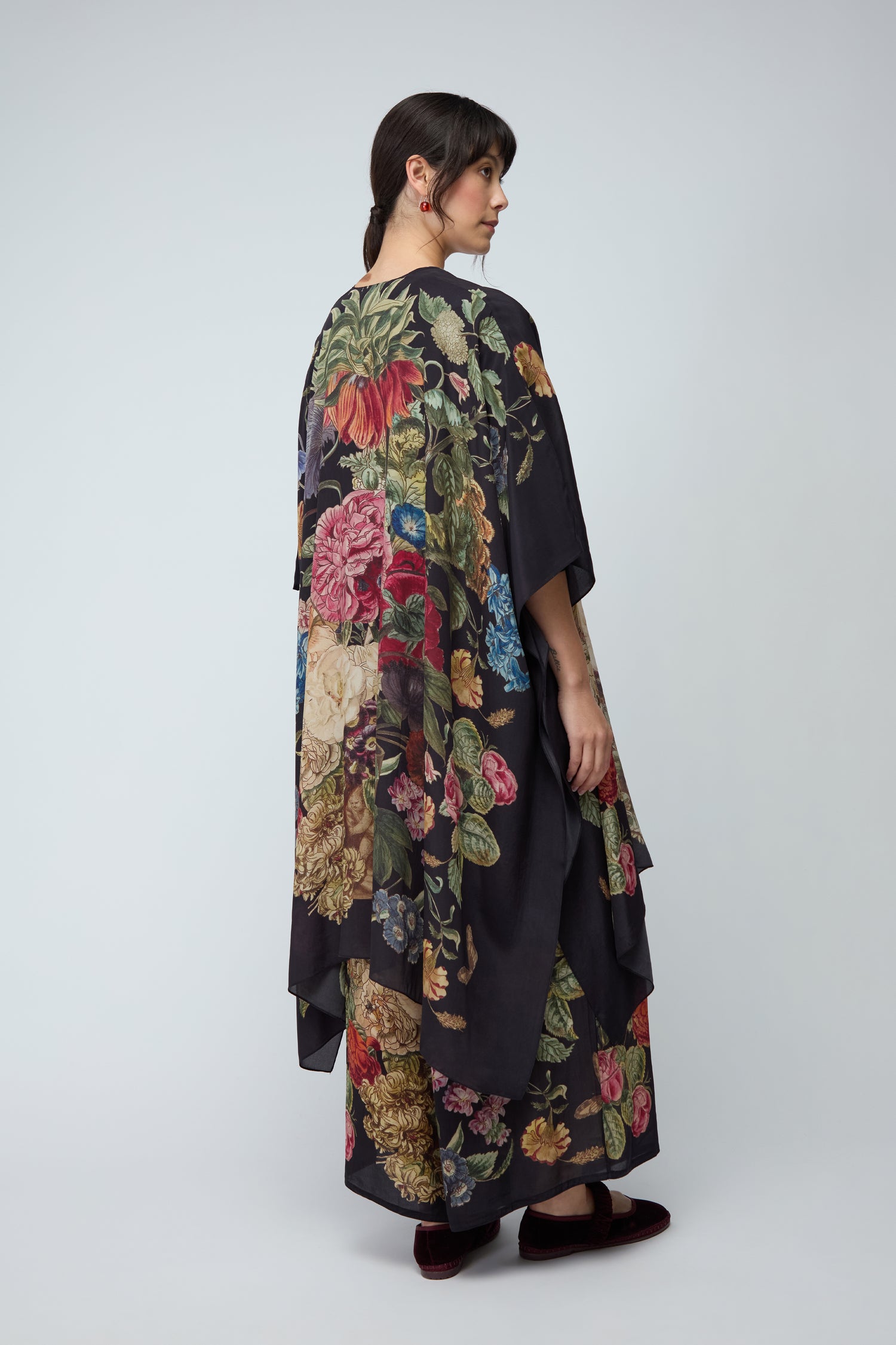 A woman with dark braided hair wears the Primula Crepe Throwover, a loose black cover-up with a primula motif and dark shoes, standing against a plain light grey background.