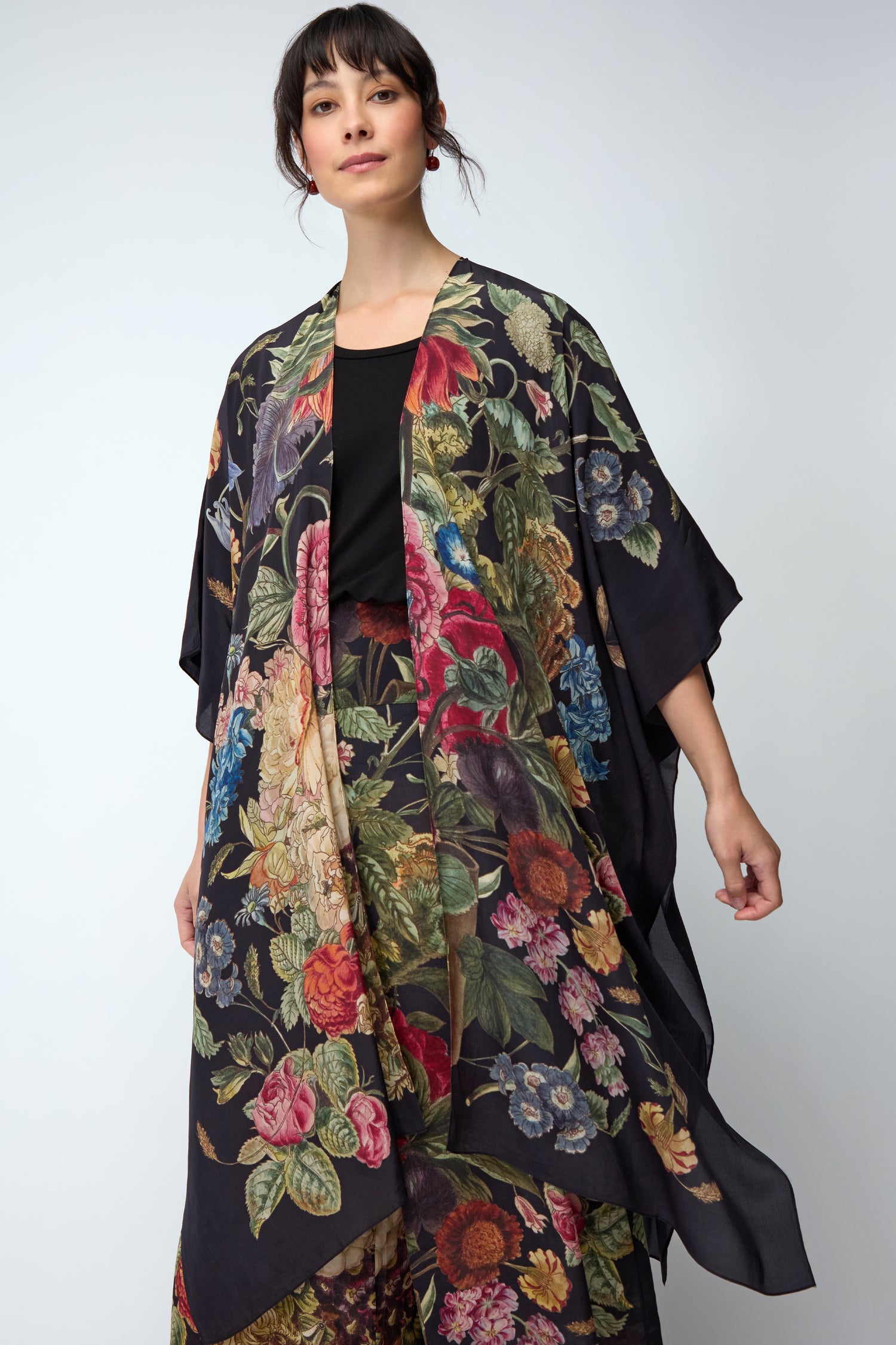 A woman wears the Primula Crepe Throwover—a black floral kimono-style garment—over a black top, standing against a plain light background.