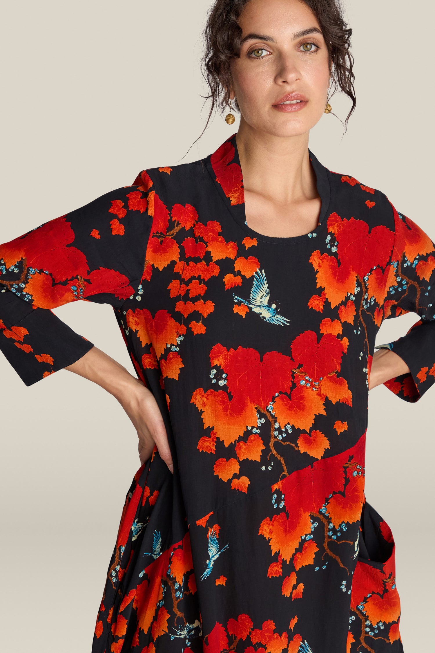 A woman wearing an Acer Print Asymmetric Dress with vibrant red and orange floral patterns and blue birds, posing with hands on hips against a plain background.