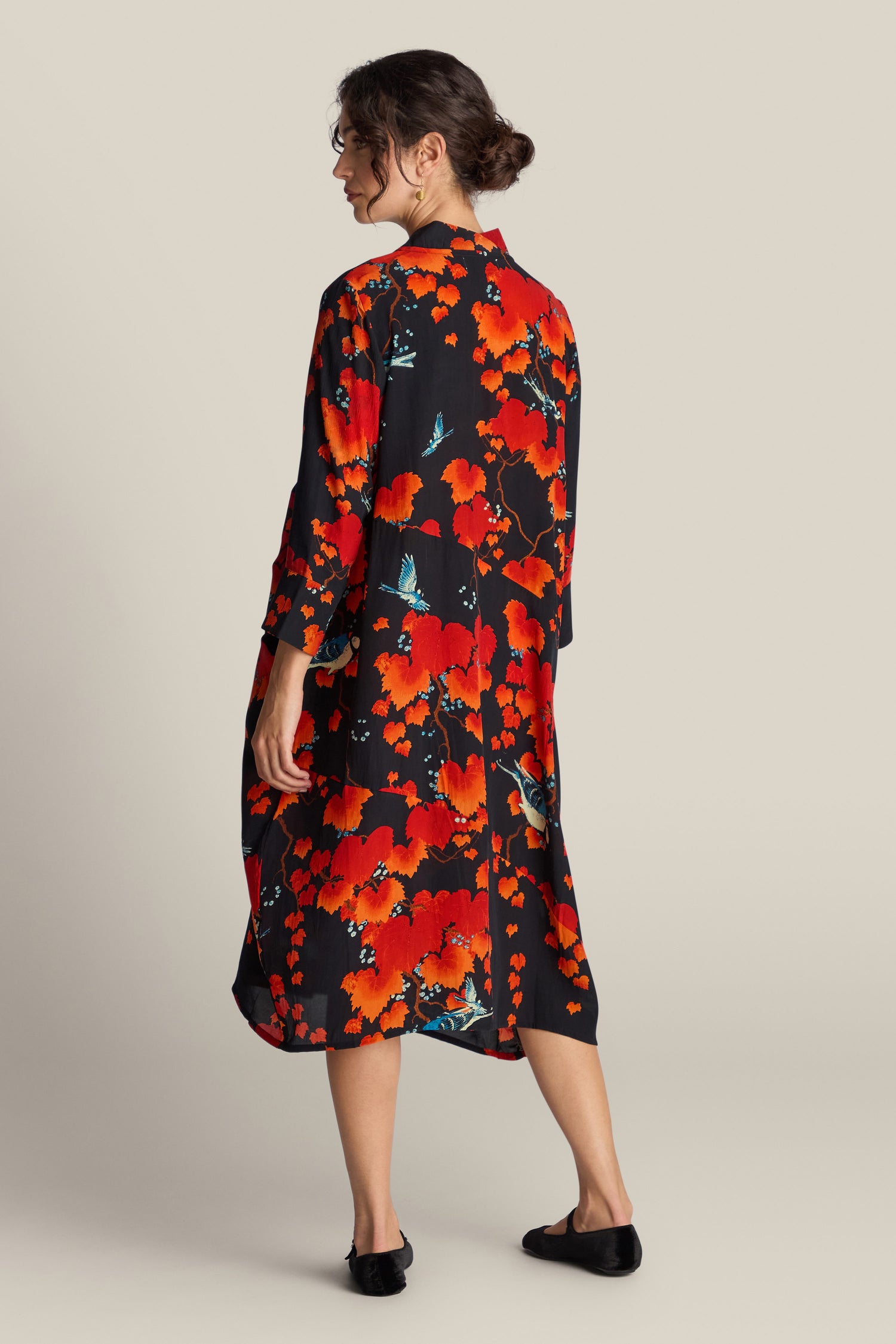 A person is standing with their back to the camera, wearing an Acer Print Asymmetric Dress—a black long dress with red and blue floral patterns—and black shoes.