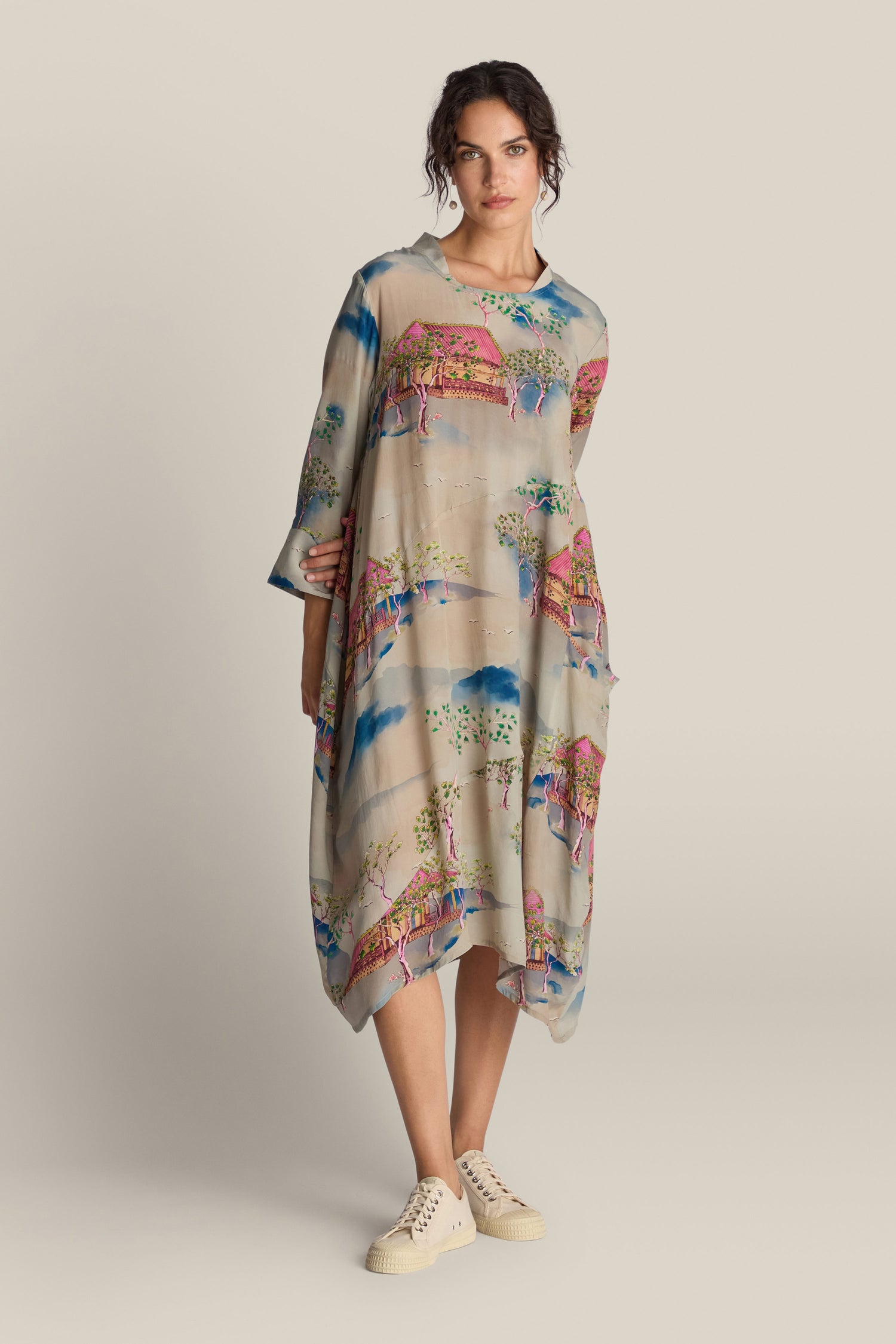 A person wearing the Landscape Asymmetric Dress, featuring vibrant designs and a knee-length hem, standing against a neutral background. They are also sporting cream-colored sneakers.