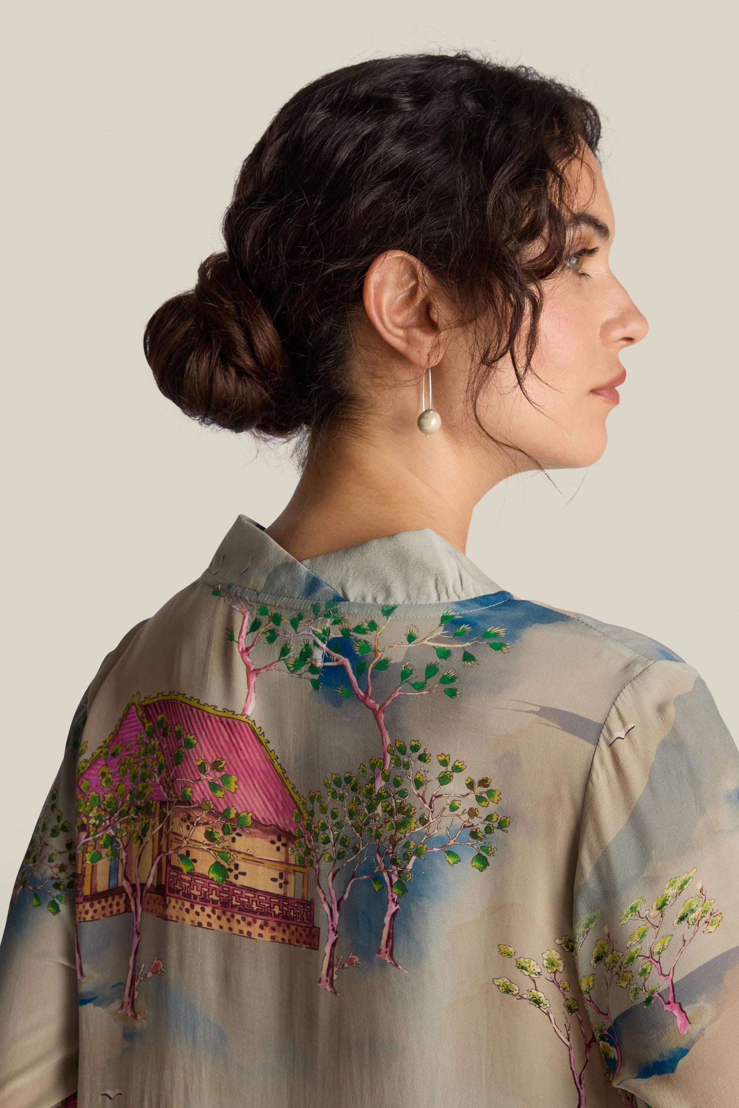 A woman with dark hair tied in a bun, wearing the Landscape Asymmetric Dress featuring an embroidered design of trees and a house, is shown in profile against a neutral background. The dress showcases a relaxed fluted neckline reminiscent of Japanese landscape watercolours.