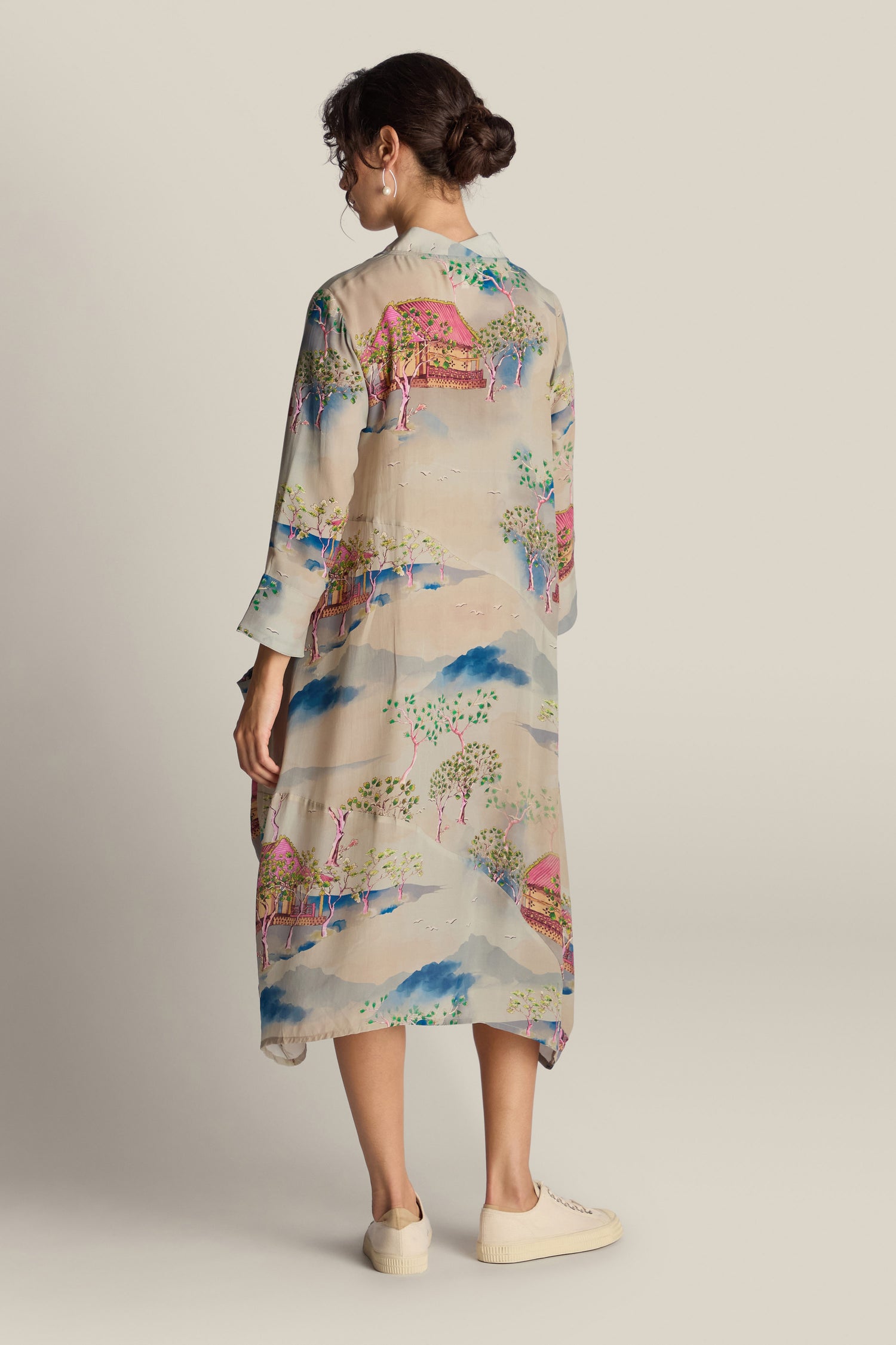 A person is wearing the Landscape Asymmetric Dress, a long, light-colored garment adorned with a vibrant Japanese landscape watercolor pattern. The dress features a relaxed fluted neckline and an asymmetric hemline. Paired with white sneakers, their hair is styled in a low bun as they look to the side against a plain beige background.