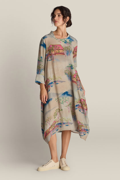 A person wearing the Landscape Asymmetric Dress, a patterned and loose-fitting dress with a nature motif and an asymmetric hem, paired with beige canvas sneakers stands against a plain background.