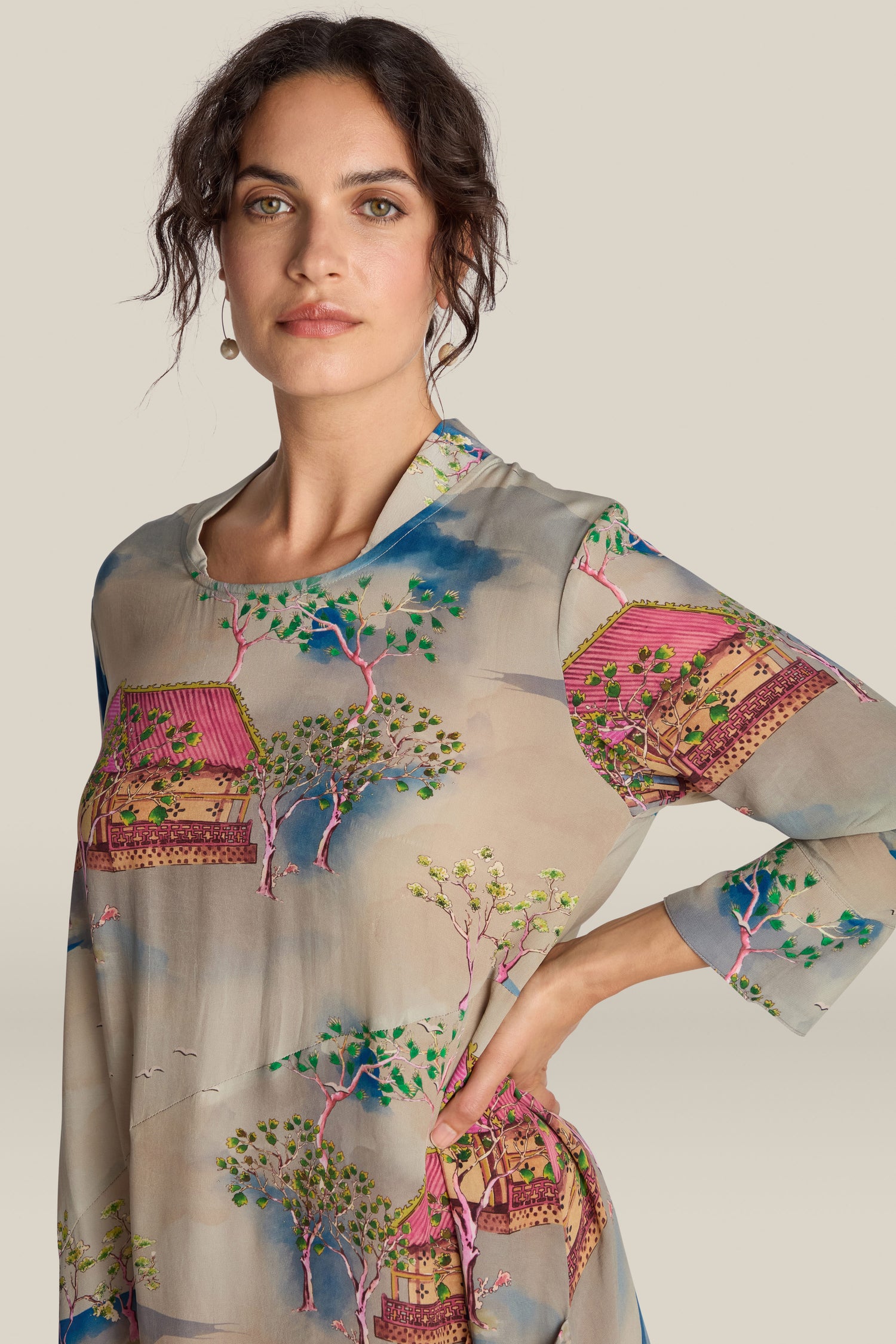 A person stands against a plain background, wearing the Landscape Asymmetric Dress, which is adorned with colorful house and tree patterns and features a relaxed fluted neckline.