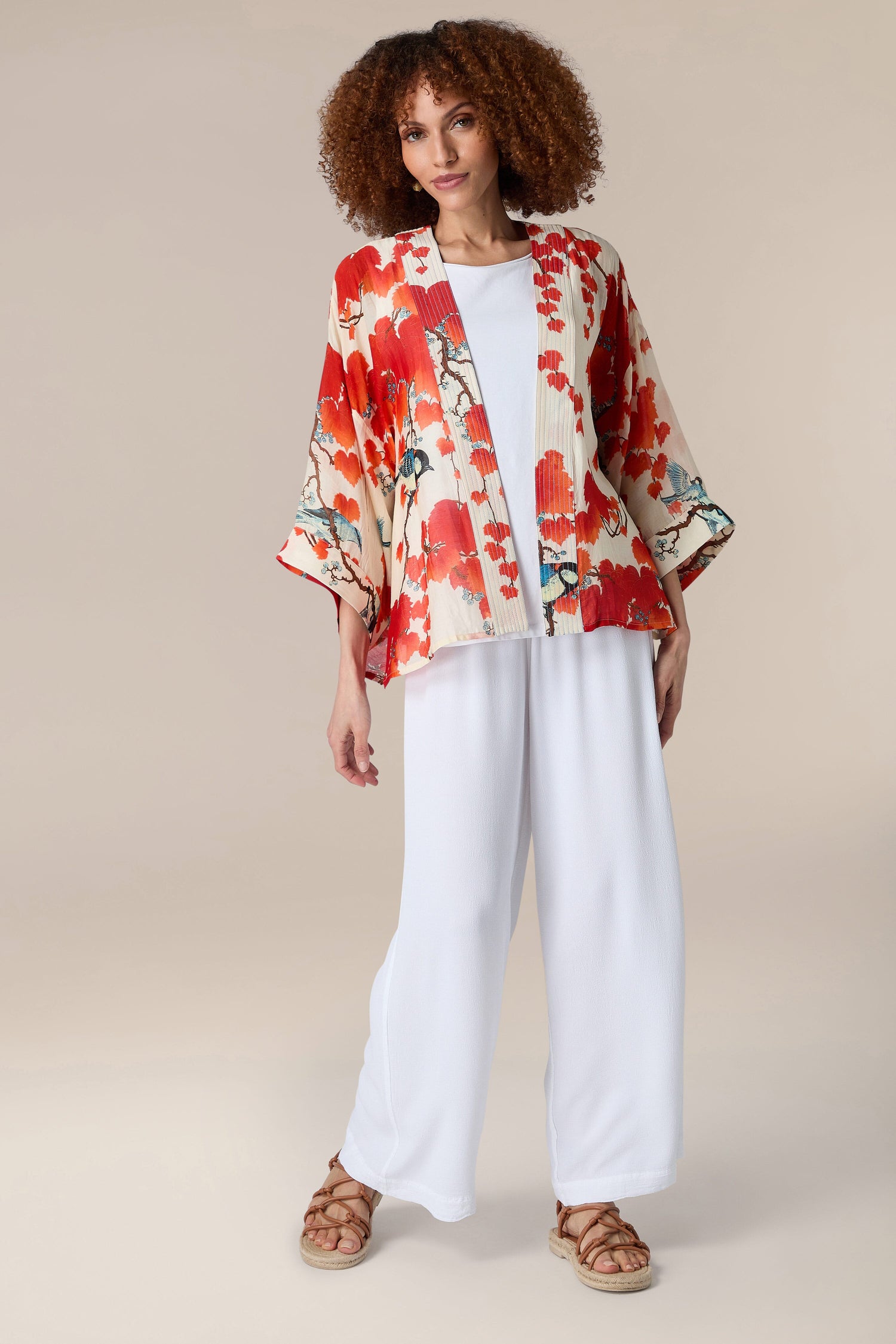 A woman in a red floral Acer Kimono and white pants walks past a Japanese maple tree.