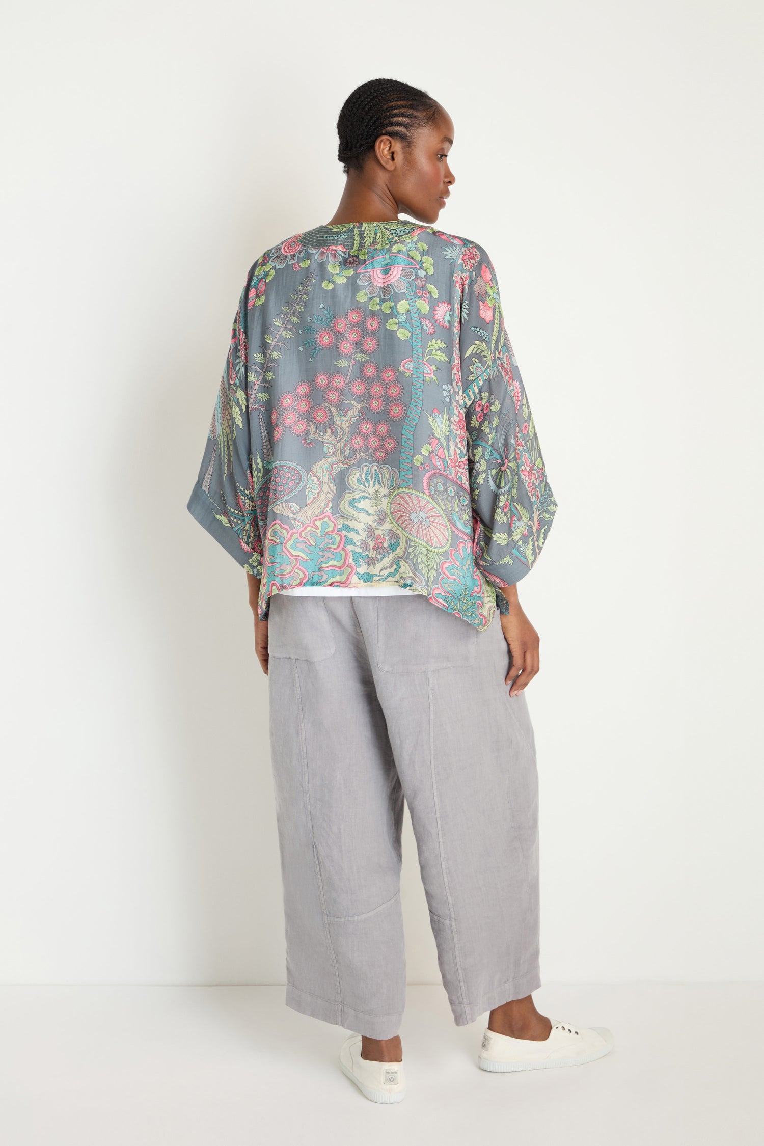 A woman stands facing away, wearing the Boho Garden Slate Print Kimono with gray wide-leg pants and white sneakers against a plain white background.