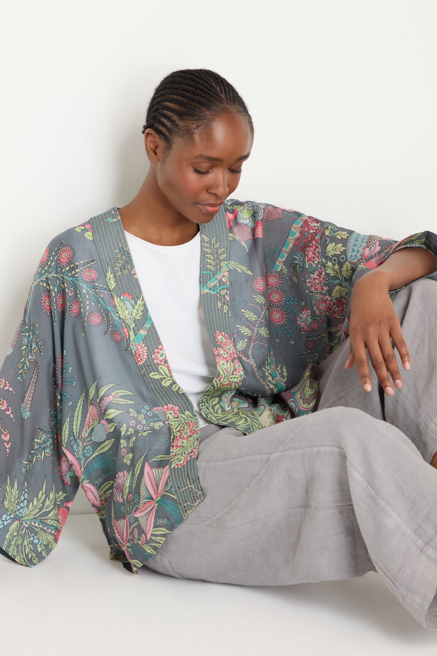 A woman sits against a white background, wearing the Boho Garden Slate Print Kimono with an archive-inspired pattern over a white shirt and light grey pants, looking down with a relaxed expression.