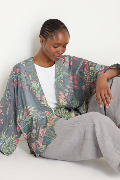 A woman sits against a white background, wearing the Boho Garden Slate Print Kimono with an archive-inspired pattern over a white shirt and light grey pants, looking down with a relaxed expression.