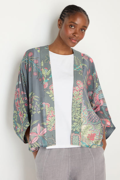 A person stands against a plain background, hands in pockets and smiling slightly, wearing a white shirt, light grey pants, and layering with the Boho Garden Slate Print Kimono featuring an archive-inspired floral pattern.