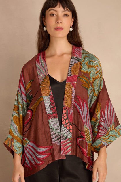 A woman wears the Crane Kimono—maroon, silky, with colorful crane and floral patterns—over a black top, standing against a beige background.