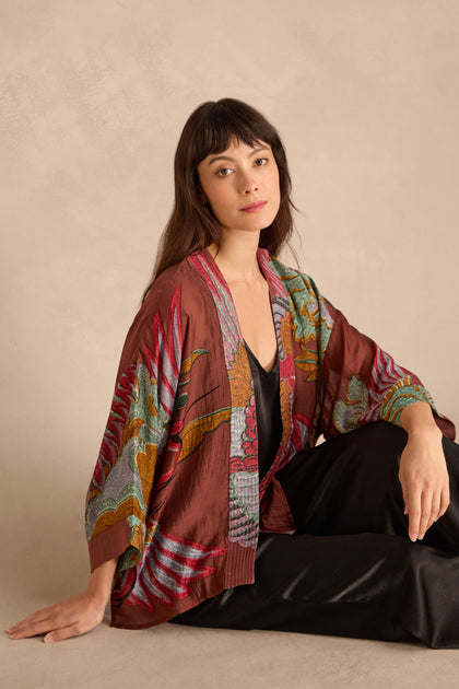 A woman with straight brown hair sits on the floor in a Crane Kimono over a black outfit, against a plain beige background.