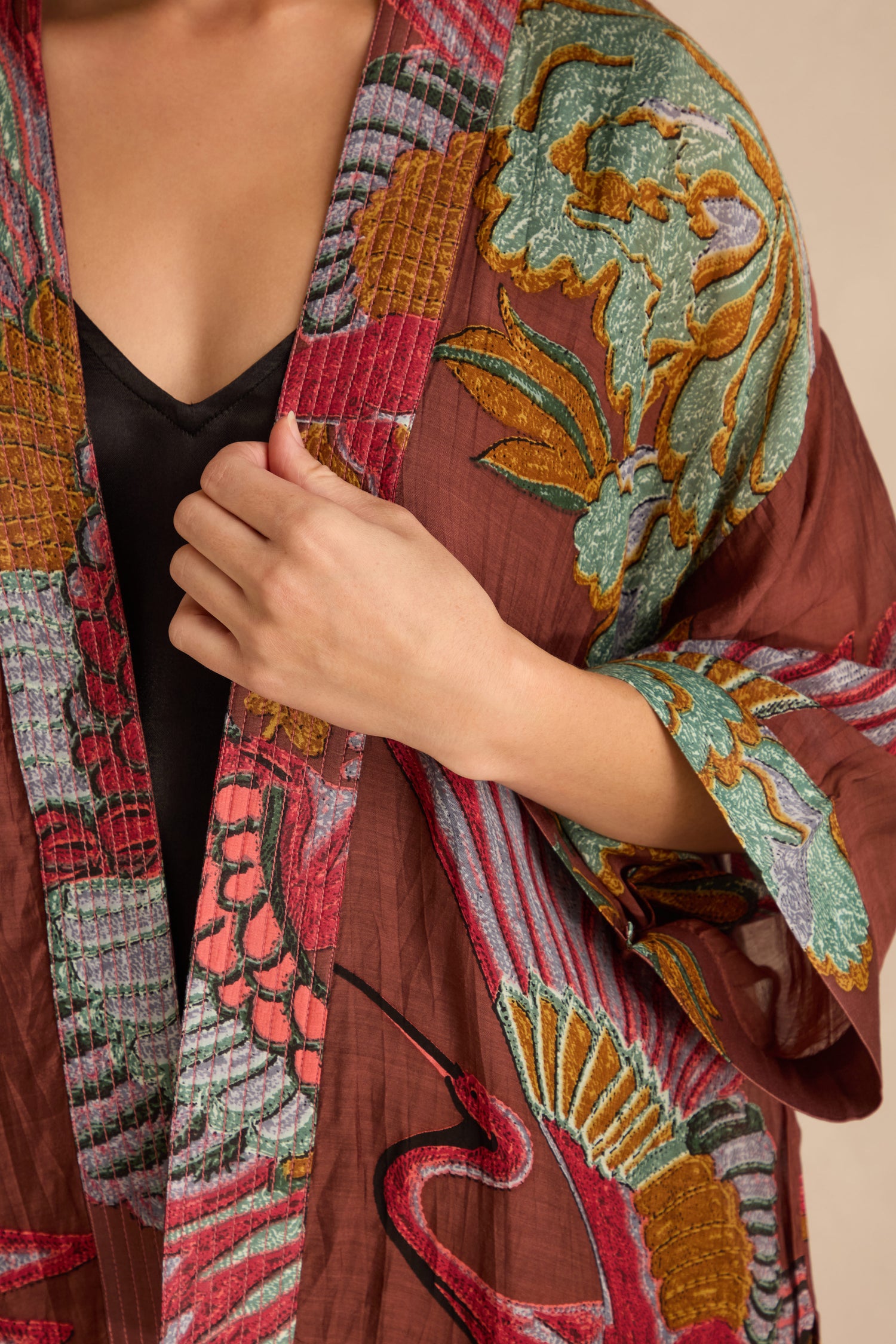 Person wearing the Crane Kimono—a silky burgundy layer with colorful embroidery—over a black top, holding the fabric with one hand.