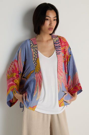 A woman stands against a plain wall wearing the Crane Slate Print Kimono over a white top and beige pants.