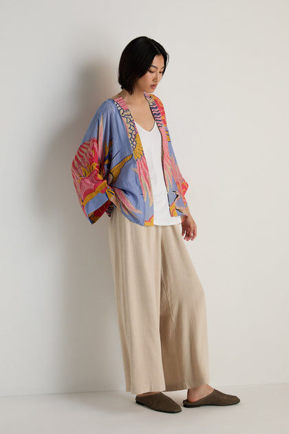 A woman stands against a plain wall wearing a Crane Slate Print Kimono, a white top, wide beige pants, and brown slippers, looking down with her hands at her sides.