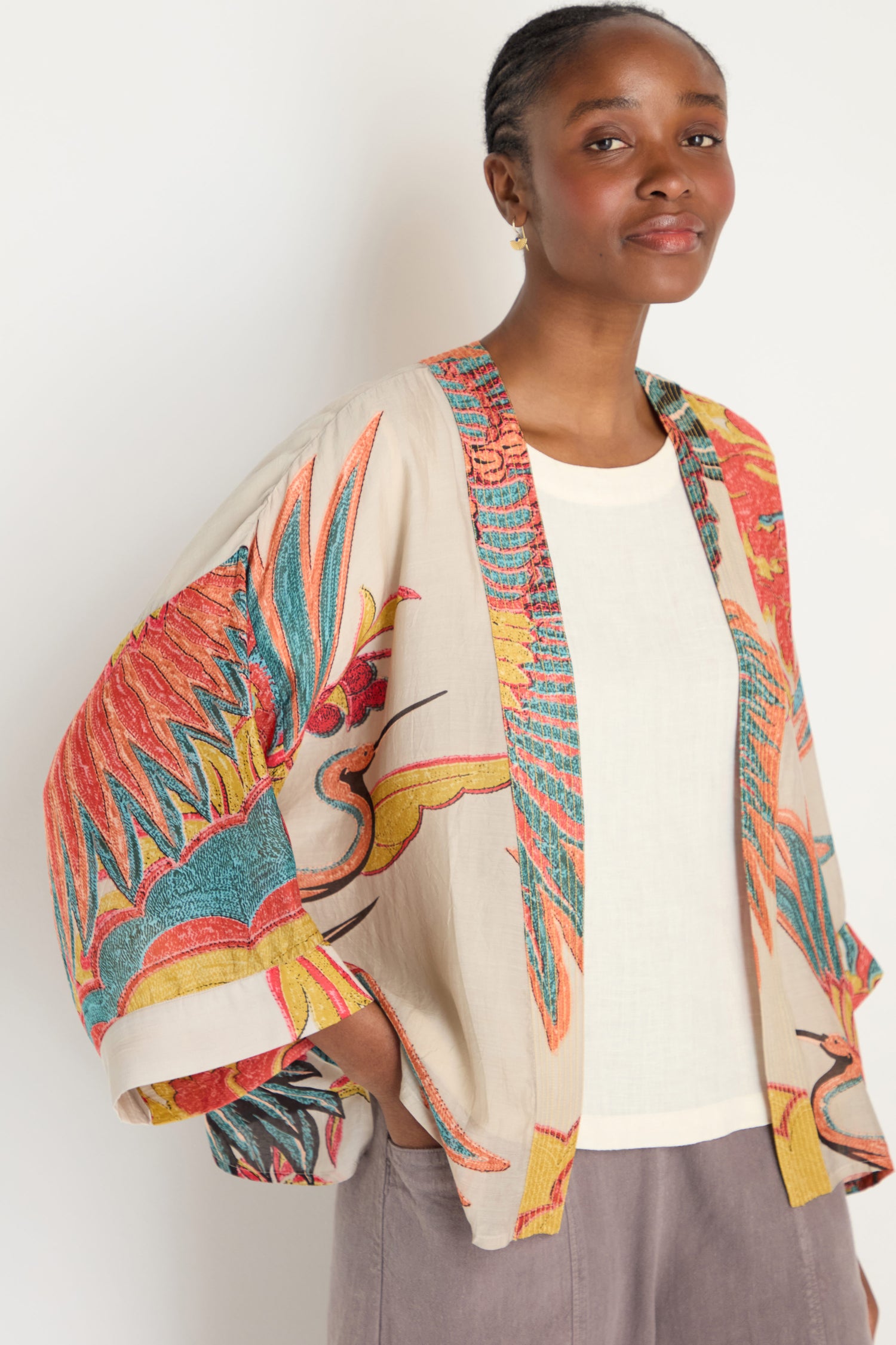 A woman stands against a plain background wearing the Crane Stone Print Kimono, a loose jacket adorned with colorful crane and feather designs—an elegant nod to timeless prints.