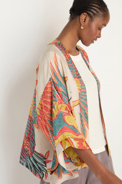 A woman in profile wears a colorful Crane Stone Print Kimono over a white top and gray pants, standing against a plain light background.