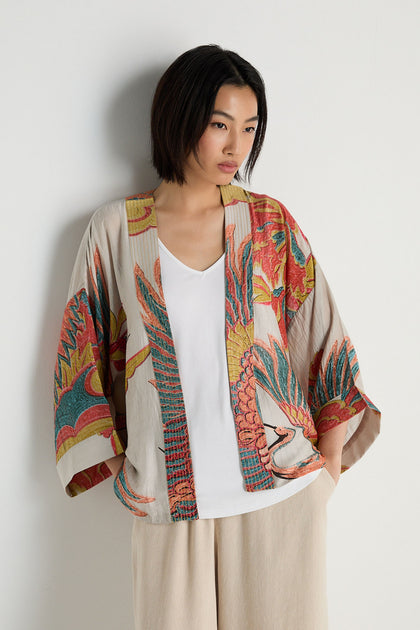 Woman with short black hair wears a Crane Stone Print Kimono over a white top and beige pants, standing against a plain light background.
