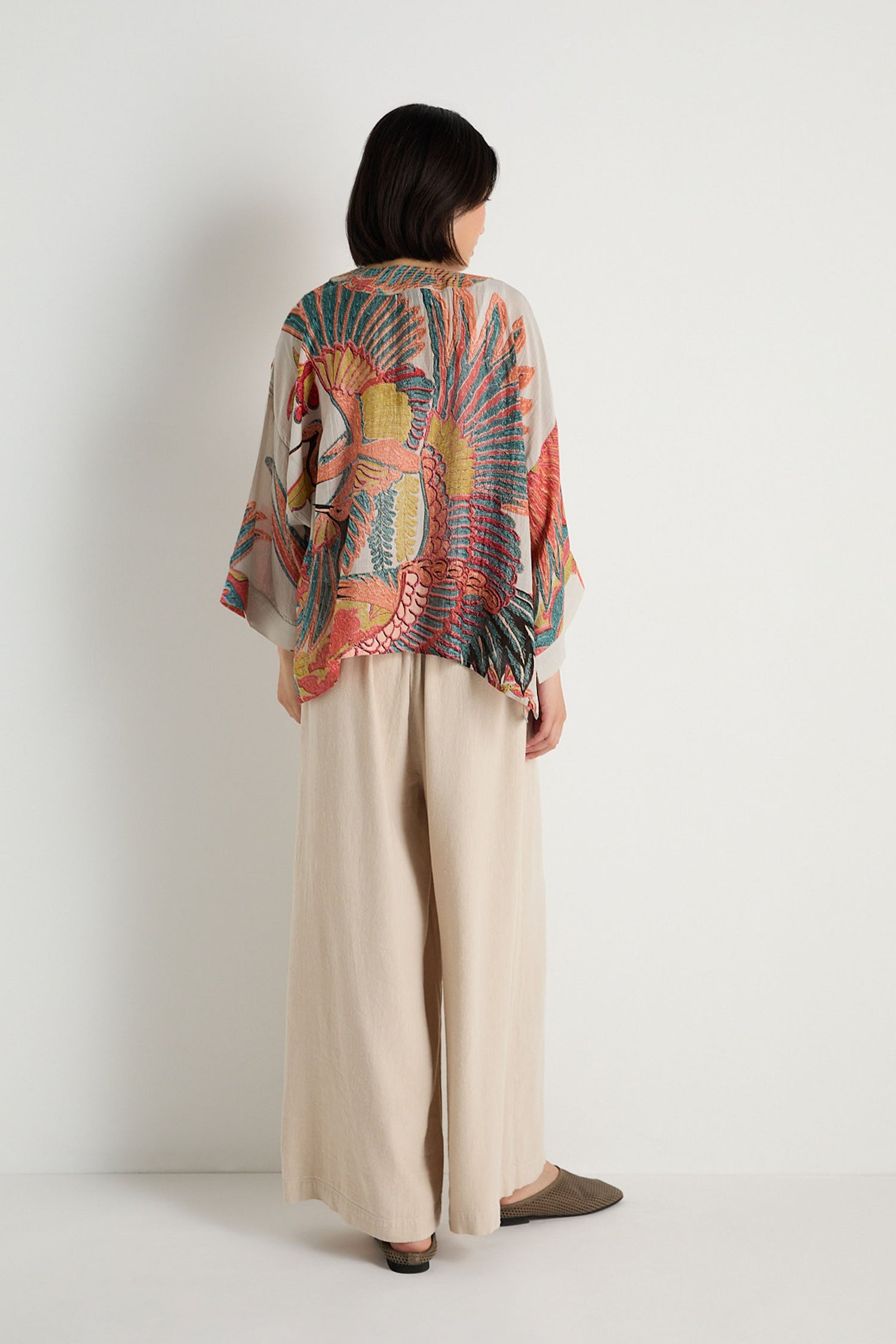 A person stands facing a white wall, wearing wide-legged beige pants, brown shoes, and a loose Crane Stone Print Kimono by One Hundred Stars featuring a bold floral and bird pattern on the back.