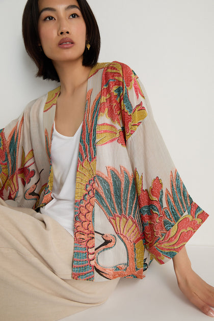 A woman in a white top and beige pants sits against a white wall, draped in the Crane Stone Print Kimono, featuring colorful floral and crane patterns.
