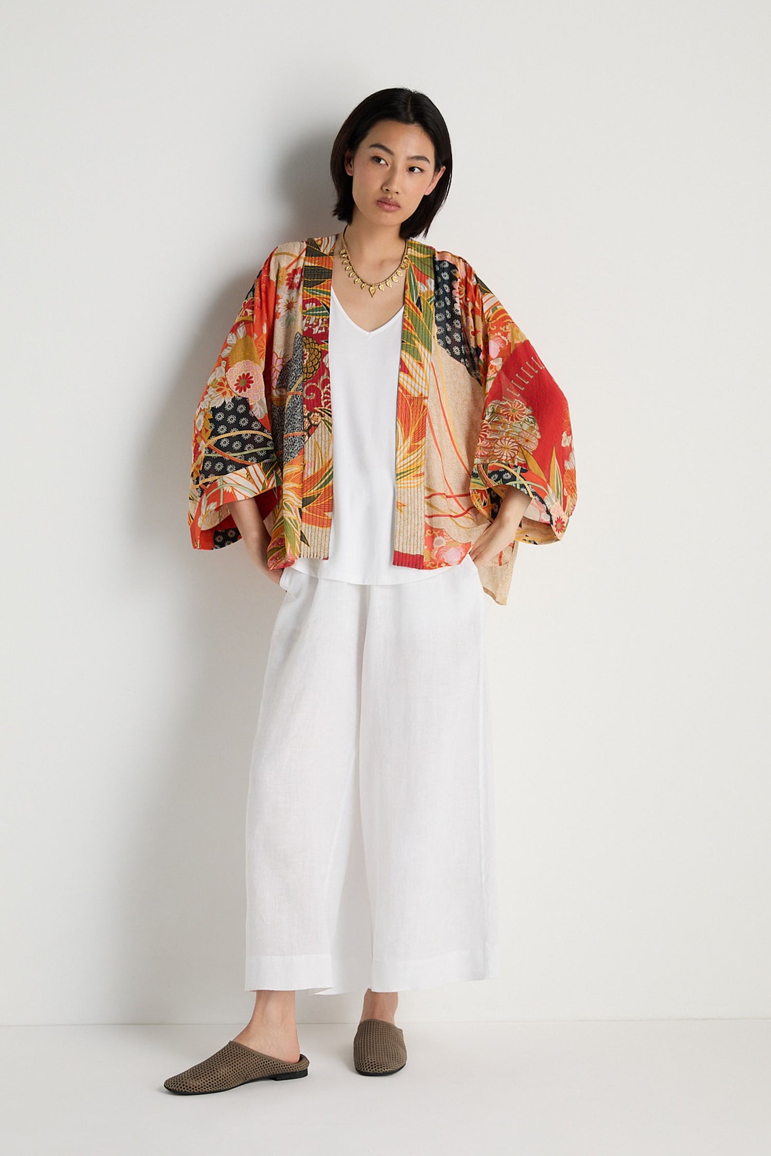 A person stands against a white wall wearing the Fans Kimono, a white top, wide white pants, brown shoes, and a gold necklace.