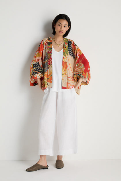 A person stands against a white wall wearing the Fans Kimono, a white top, wide white pants, brown shoes, and a gold necklace.