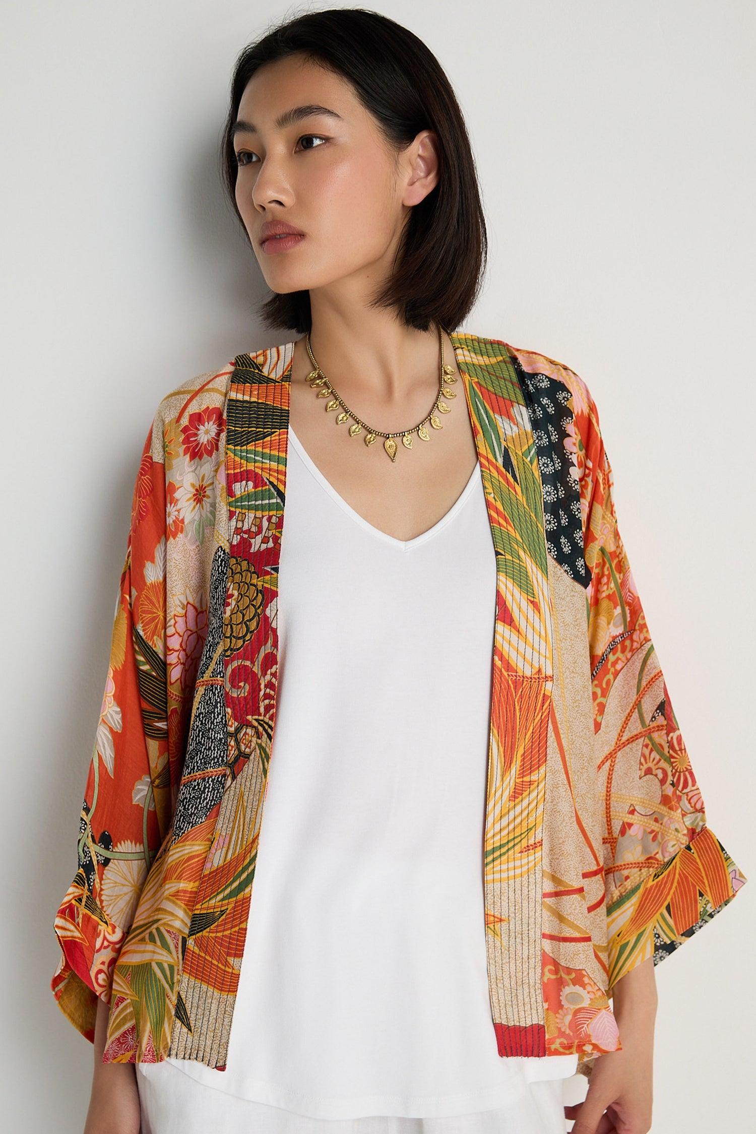 A woman wearing the Fans Kimono—a colorful fan print jacket in modal and cupro blend—over a white top stands against a plain light background, gazing to the side.