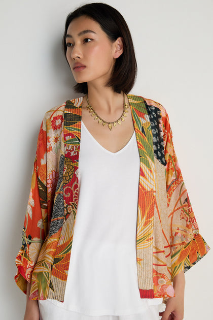 A woman wearing the Fans Kimono—a colorful fan print jacket in modal and cupro blend—over a white top stands against a plain light background, gazing to the side.