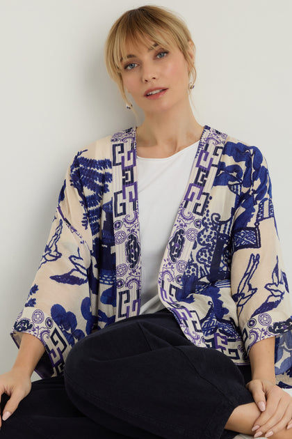 A blonde woman sits against a white wall, wearing a white shirt, black pants, and the Willow Print Kimono in blue-and-white modal and cupro.