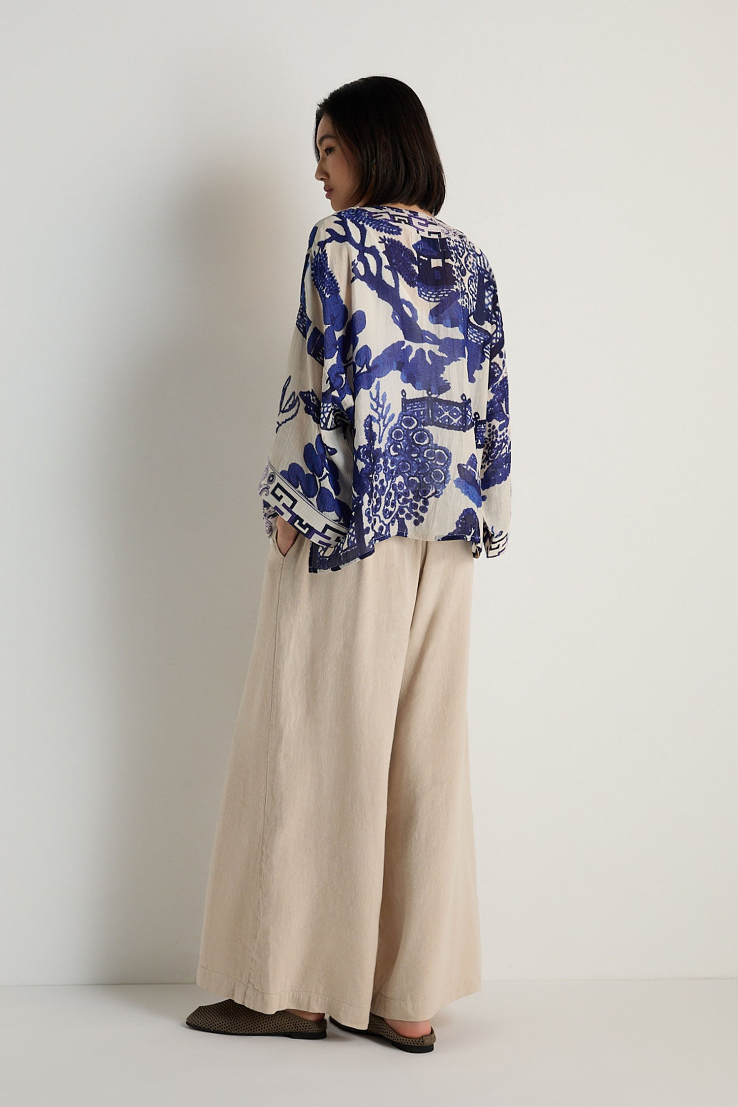 A person stands facing away, wearing the Willow Print Kimono in a blue-and-white motif, paired with wide beige pants and dark flat shoes against a plain white background.