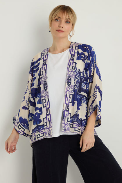 A person stands against a plain light background wearing a white shirt, dark pants, and the Willow Print Kimono, featuring blue Asian-inspired patterns and crafted from modal and cupro.