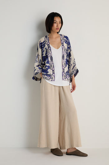 A woman stands against a plain wall wearing a white top, wide beige pants, and the Willow Print Kimono with a blue-and-white motif, styled with brown slippers for a relaxed look.