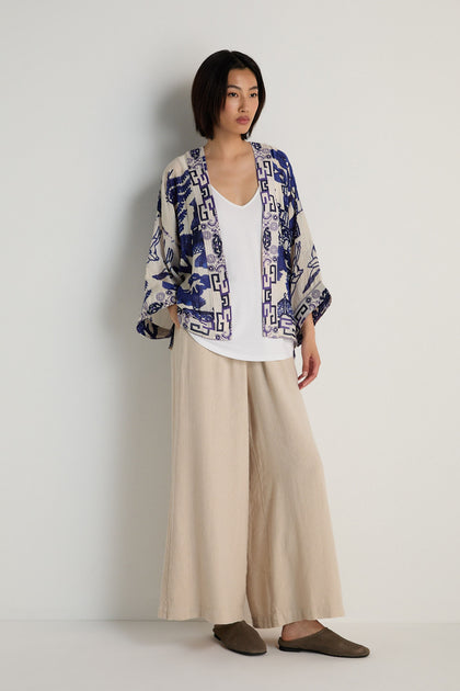 A woman stands against a plain wall wearing a white top, wide beige pants, and the Willow Print Kimono with a blue-and-white motif, styled with brown slippers for a relaxed look.