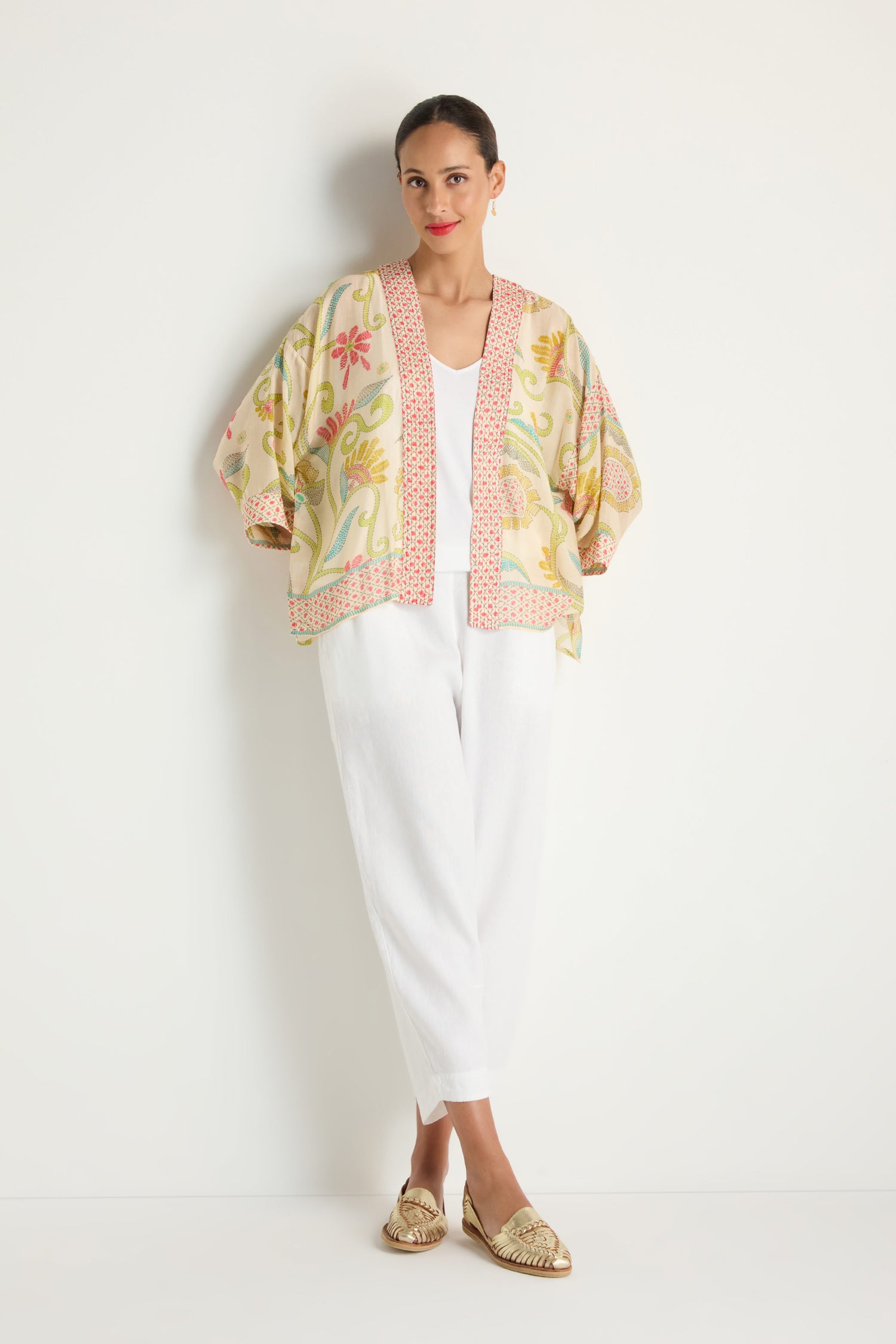 Woman stands against a white wall, wearing the Kantha Lime Kimono with white pants, a white top, and gold woven loafers.