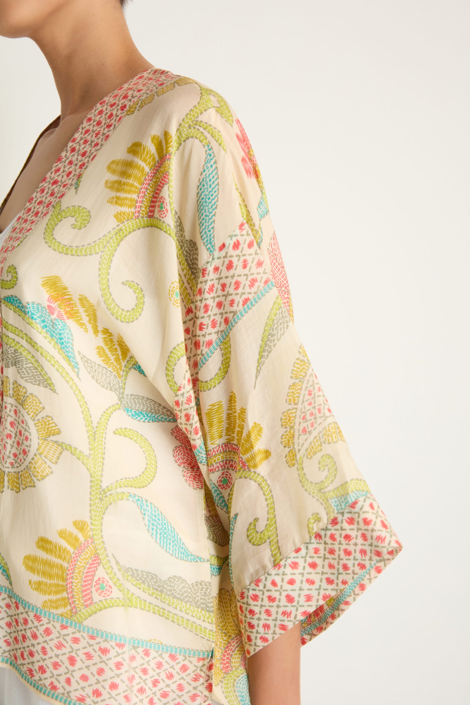 A person wearing the Kantha Lime Kimono, crafted from lightweight woven cloth in a lime ecru palette with yellow, pink, green, and blue floral and geometric designs, shown from shoulder to mid-arm.