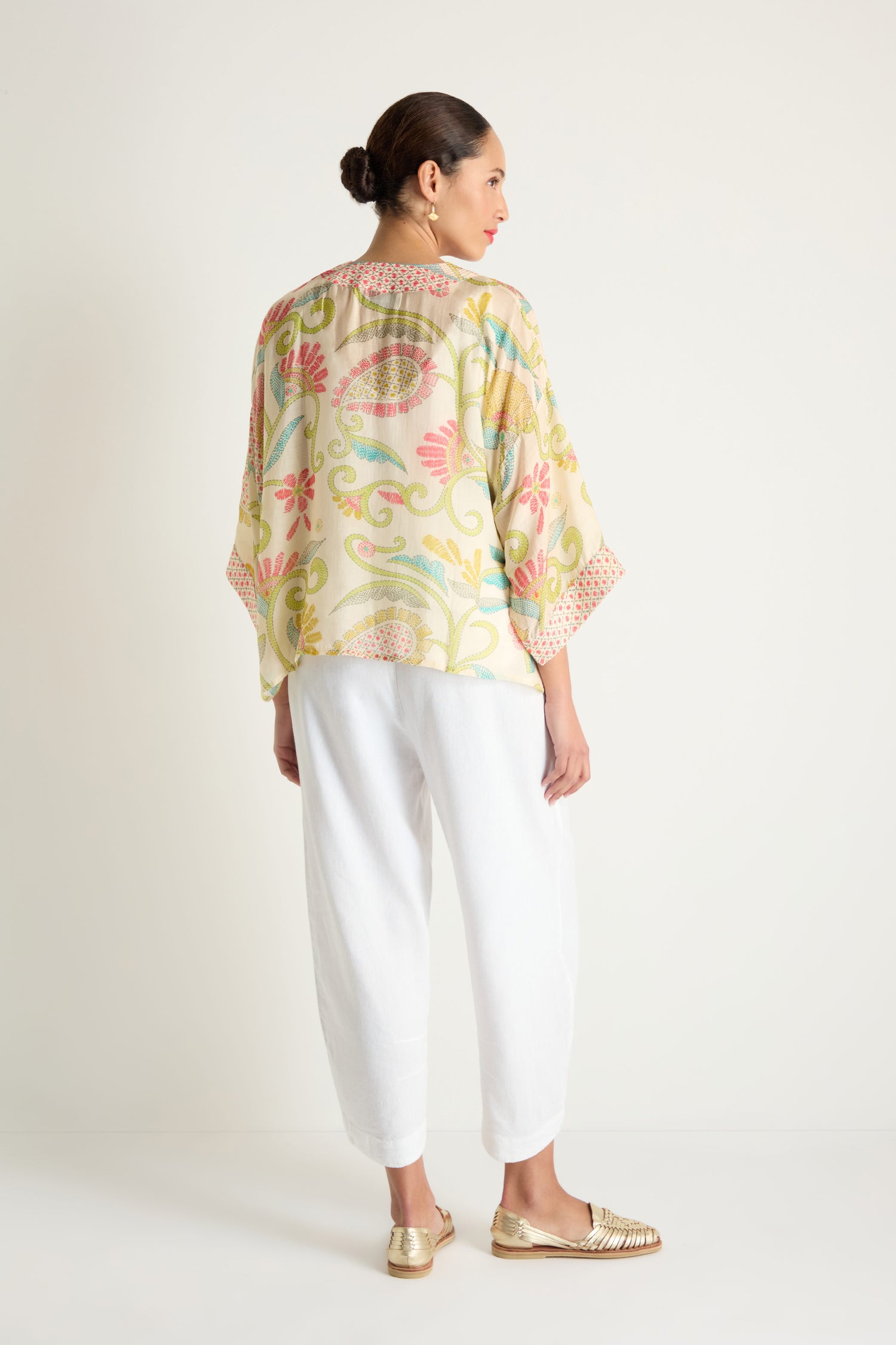 A woman faces away from the camera against a plain background, wearing the Kantha Lime Kimono in pastel lime and ecru tones, styled loosely with white pants and gold shoes.